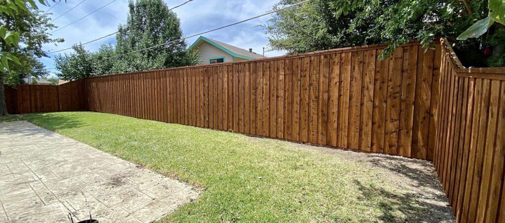 A tall wooden privacy fence runs along the length of a grassy backyard, bordered on one side by a concrete walkway. Trees and part of a house with a green-trimmed roof are visible in the background, prompting thoughts like, "Should I Install Fence Poles on the Inside or Outside?.