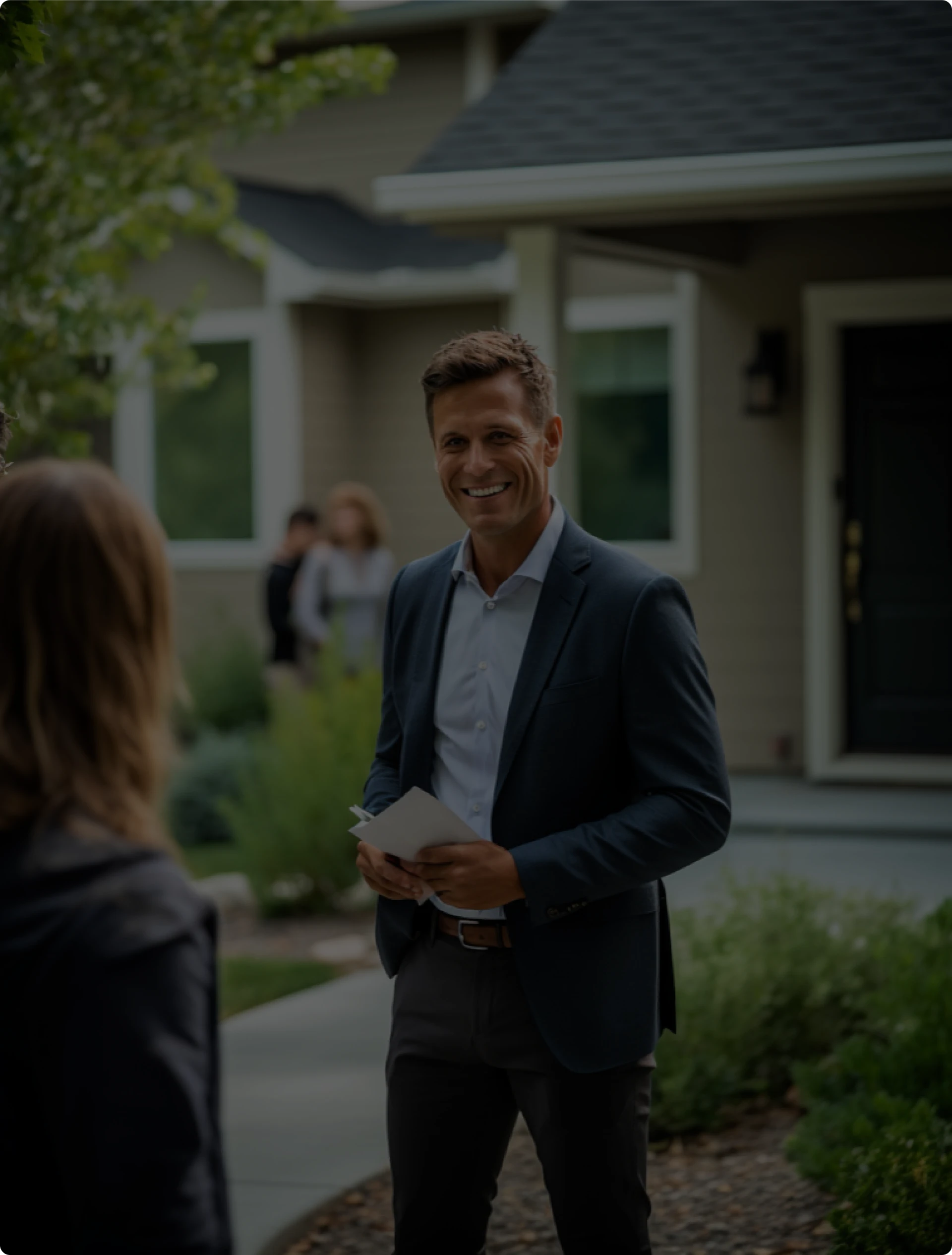 A man in a suit stands outside a house, smiling and holding papers, while facing a woman. Two people are visible in the background near the entrance. The setting appears to be a residential neighborhood.