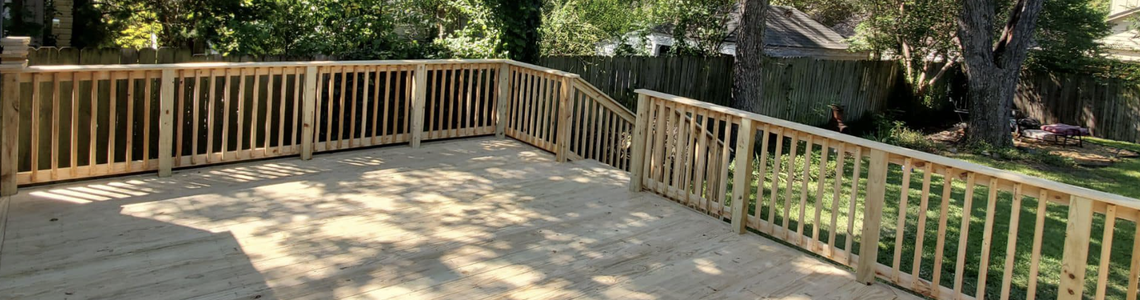 A newly built wooden deck with a railing surrounds a spacious, sunlit area. Stairs lead down to a grassy Dallas backyard with trees, custom wood fence design, and some outdoor furniture in the distance.