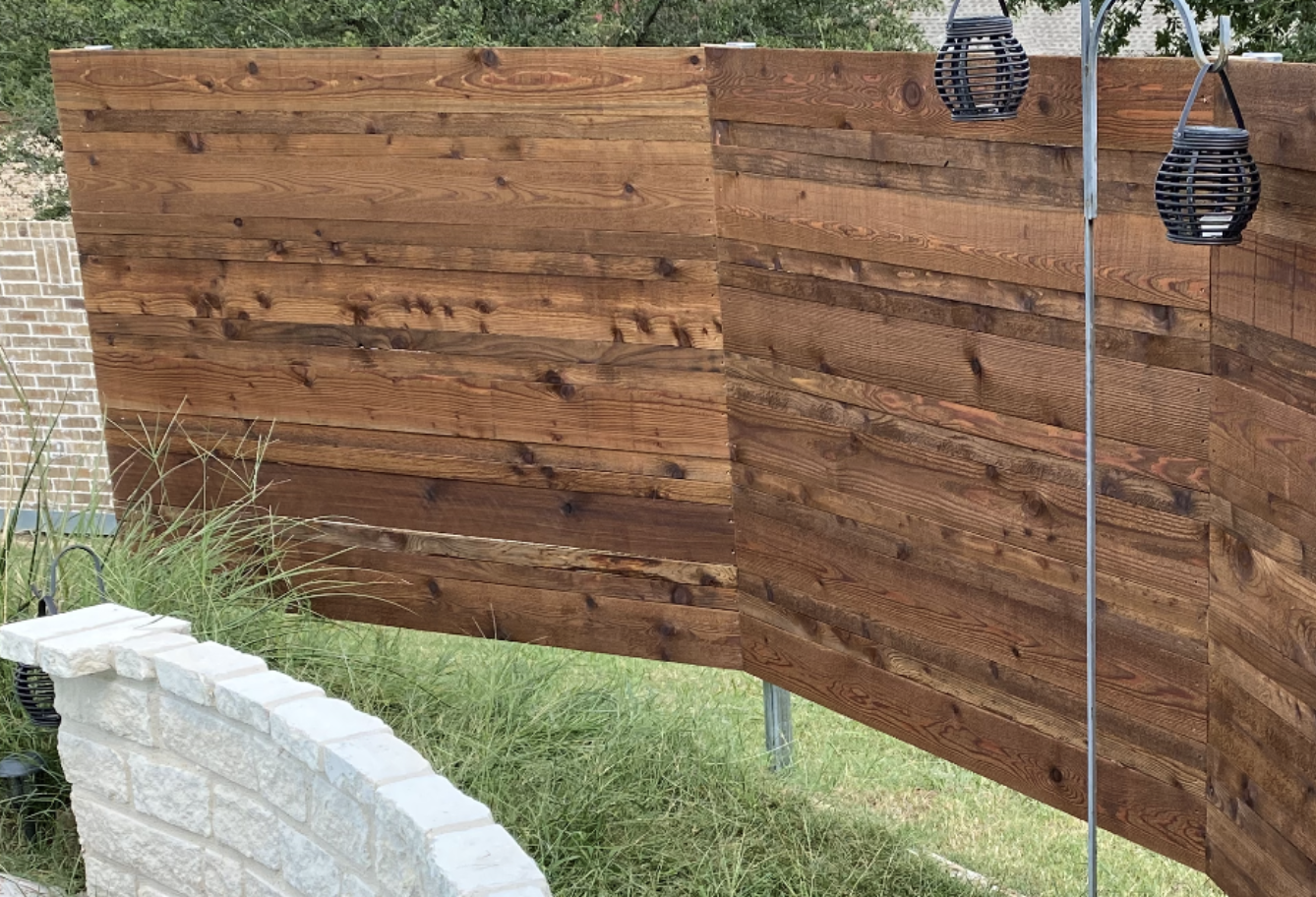 Two wooden privacy fence panels, showcasing custom wood fence Dallas style, stand at an angle in a backyard. Ornamental lanterns hang from poles nearby, and tall grass grows in front of a light-colored stone retaining wall.