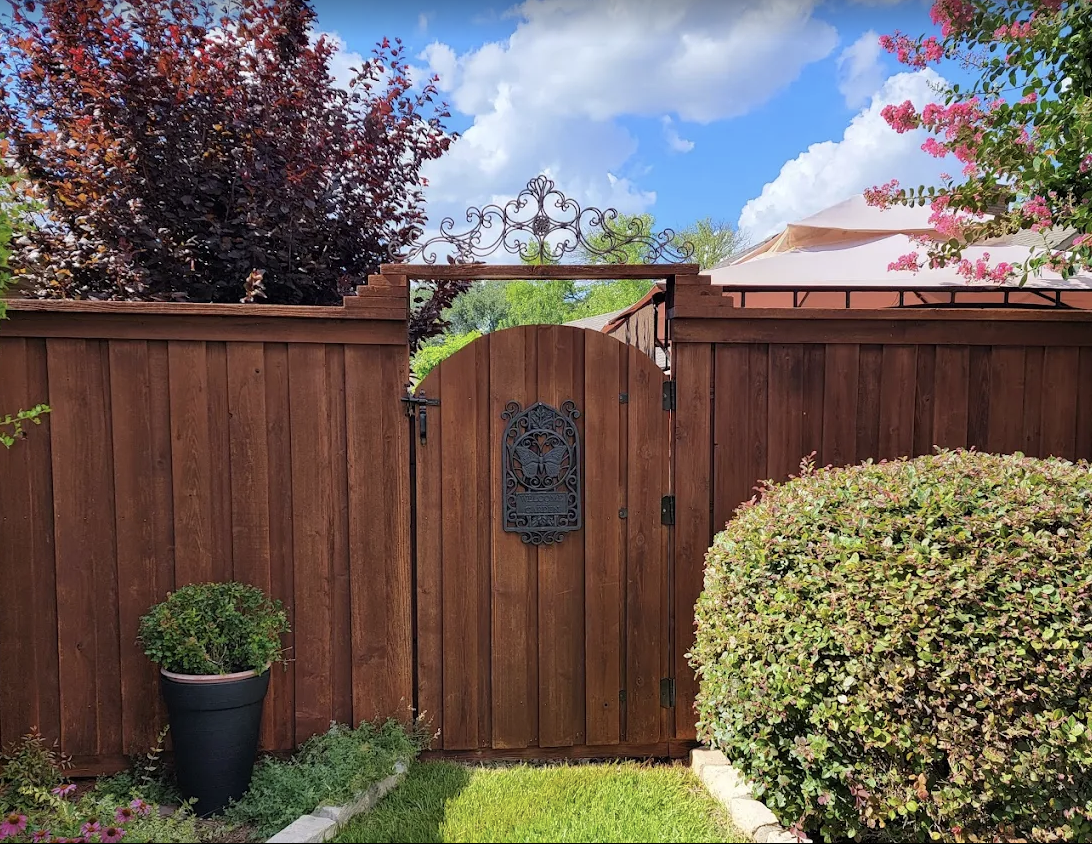 A wooden garden fence made from the best wood for a fence, with a closed arched gate featuring ironwork decorations. A potted plant sits on the left and a large shrub on the right under a blue sky dotted with scattered clouds.