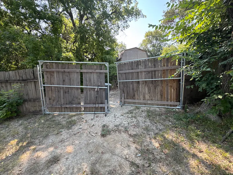 A wooden double gate with a metal frame is unevenly installed on a dirt pathway, hinting at the need for wood fence repair. The surrounding area features trees, bushes, and remnants of an old fence that could benefit from regular wood fence maintenance.