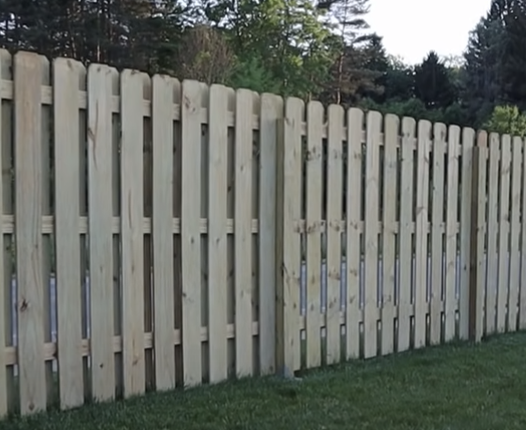 A wooden picket fence with vertical planks stands on a grassy lawn, showcasing a classic wood fence design, with trees and greenery visible in the background—a perfect inspiration for your Custom Wood Fence Dallas project.