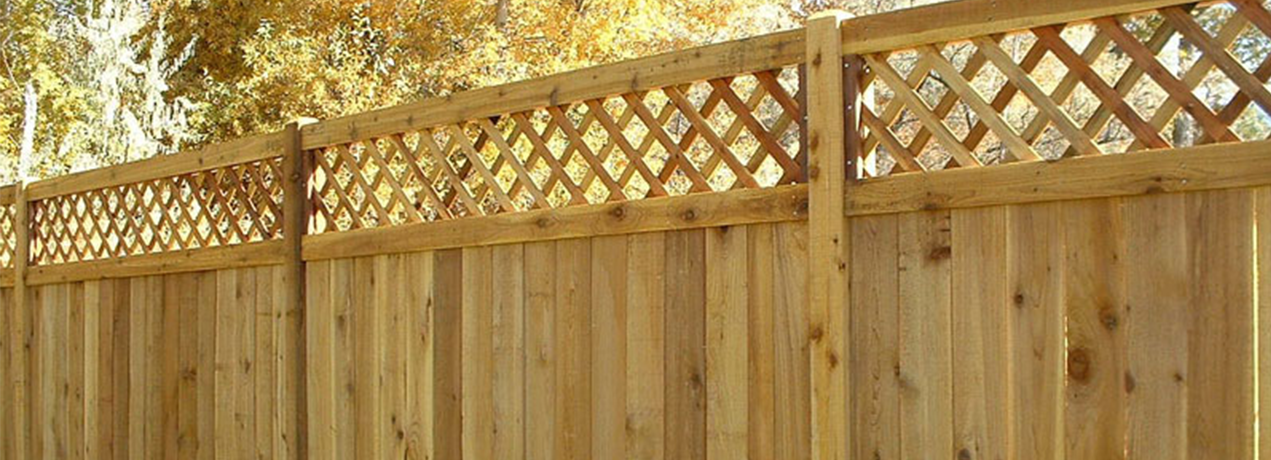 A custom wood fence in Dallas with vertical planks and a decorative lattice top runs horizontally across the image. Trees with autumn-colored leaves provide a scenic backdrop, highlighting this elegant wood fence design.