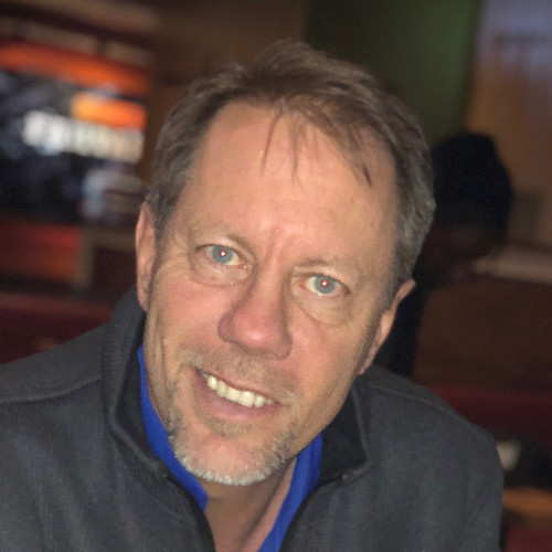 A middle-aged man with light skin, short brown hair, and a light beard is smiling at the camera. He is wearing a dark jacket over a blue collared shirt. The background is blurred.