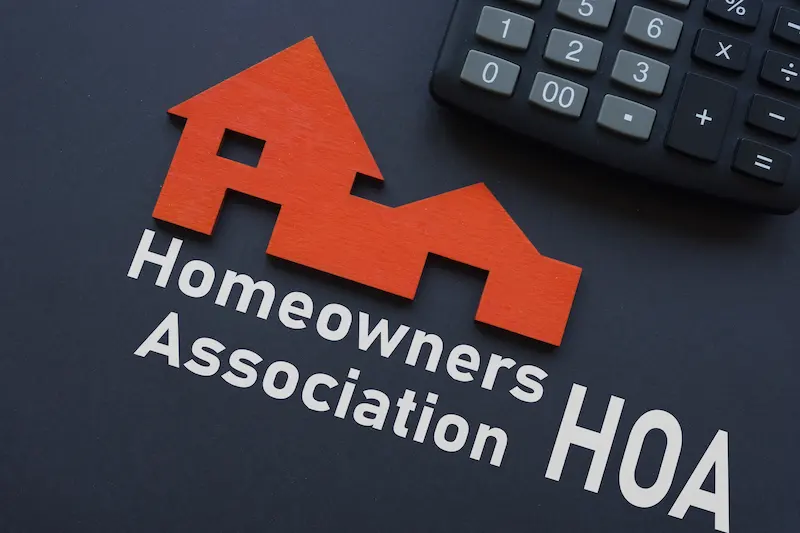 A red house-shaped logo beside the words Homeowners Association HOA appears on a black surface, with part of a gray calculator in the top right corner—symbolizing how HOAs manage fence rules and permits within communities.