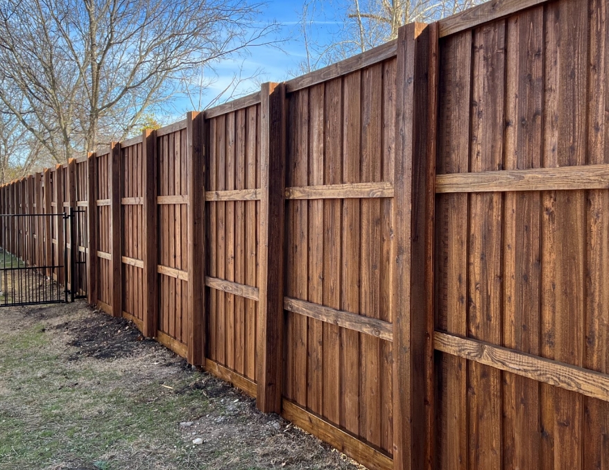 A tall wooden privacy fence with vertical planks and horizontal support beams separates a grassy yard in Mansfield TX from trees and a metal fence beyond. The wood features a rich, reddish-brown finish.