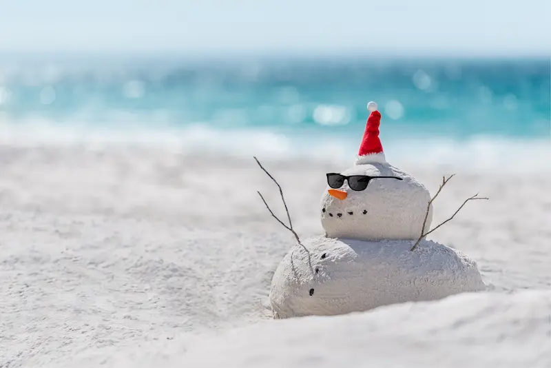 A sand snowman on a beach, wearing a red Santa hat and black sunglasses, with stick arms and a carrot nose. The ocean is blurred in the background, blending holiday cheer with thoughts of home security and package protection.