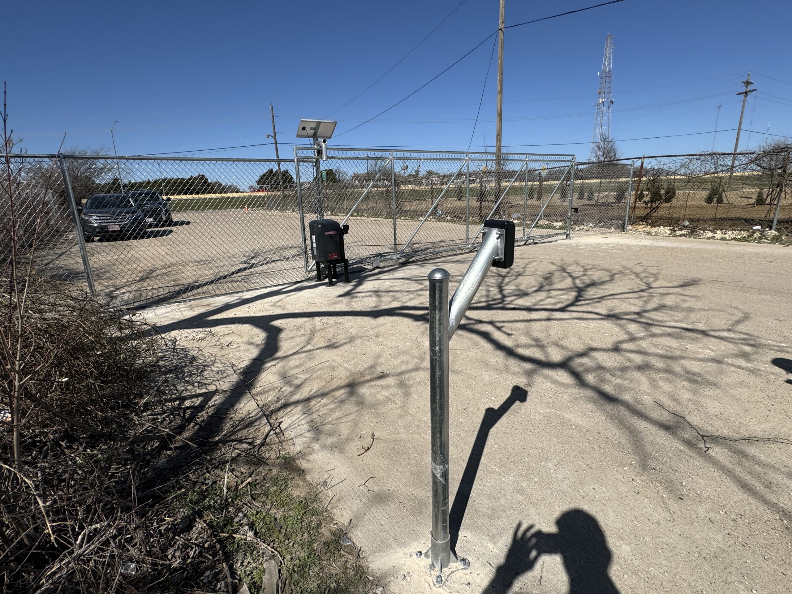 A fenced parking lot with a closed metal gate and automated keypad entry, featuring iron fence installation. A surveillance camera and utility poles stand nearby, with a few parked cars and a shadow of a person in the foreground.
