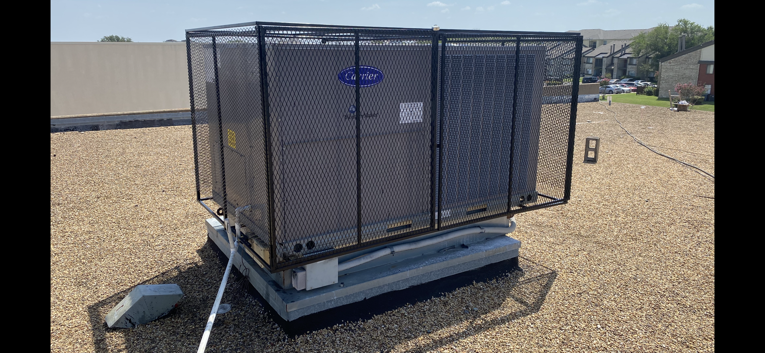 A large Carrier HVAC unit is enclosed in a metal mesh cage, reminiscent of iron fence installation, on a flat gravel rooftop. Nearby buildings and a grassy area are visible in the background under a clear sky.