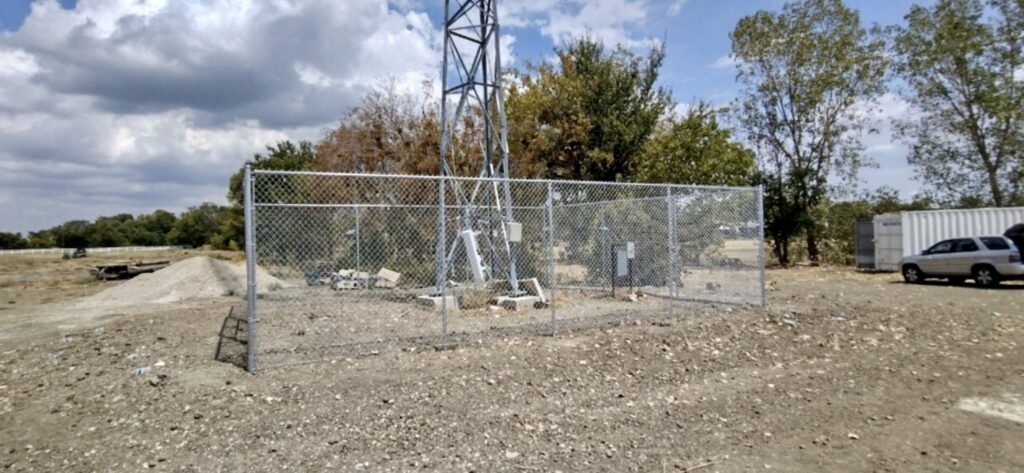 A chain-link fence encloses a small utility area with electrical equipment and a metal tower on a dirt lot, showcasing an alternative to iron fence installation. Trees, a white container, and a parked vehicle sit in the background under partly cloudy skies.