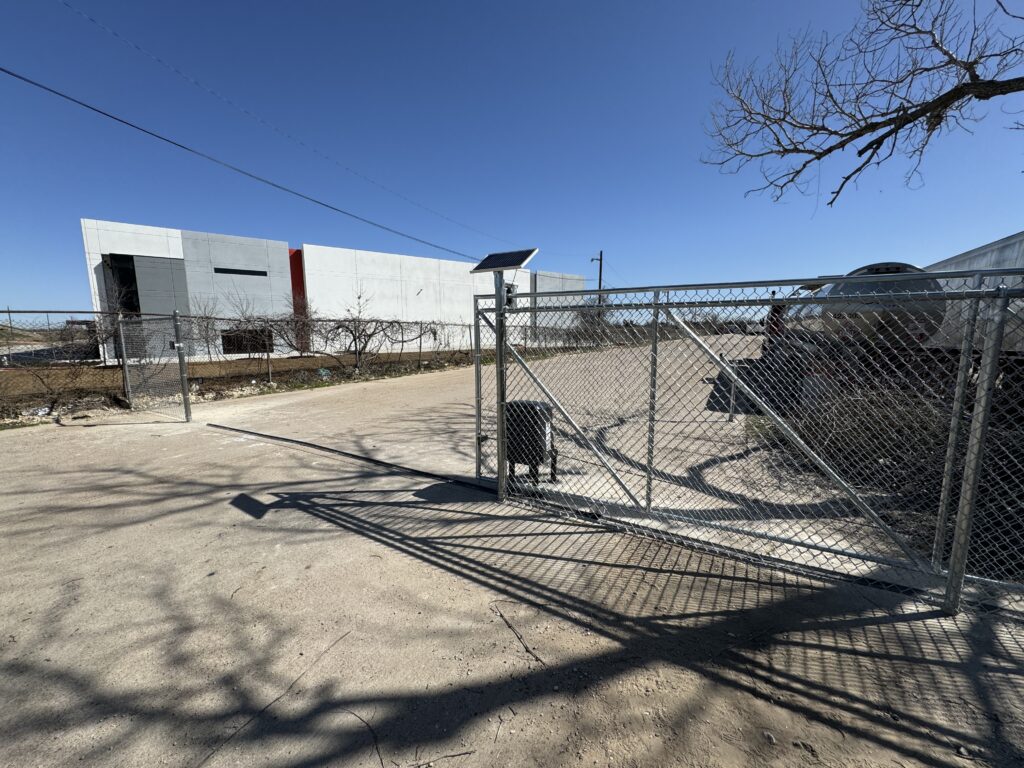 A chain-link gate, part of a recent iron fence installation, stands partially open across a paved road. A metal building rises in the background under a clear blue sky, while bare tree branches cast shadows on the ground.