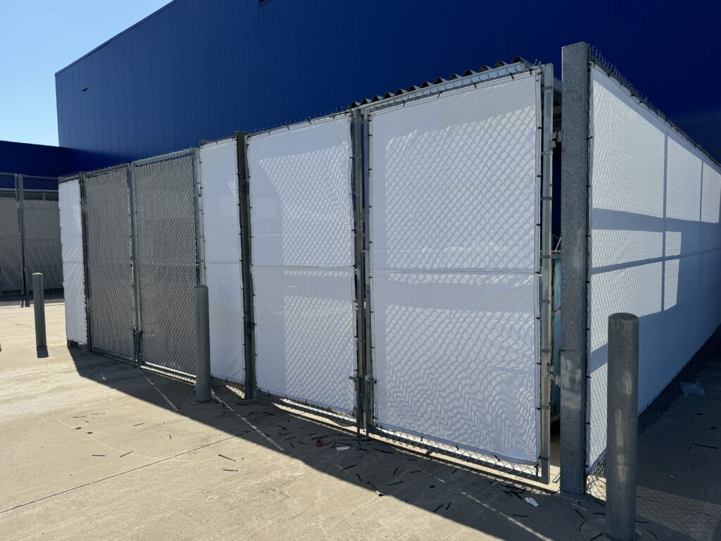 A chain-link fence, possibly serving as a temporary alternative to iron fence installation, with panels covered in white material stands next to a blue building on a concrete surface beneath a clear sky. Shadows from the fence and poles are visible on the ground.