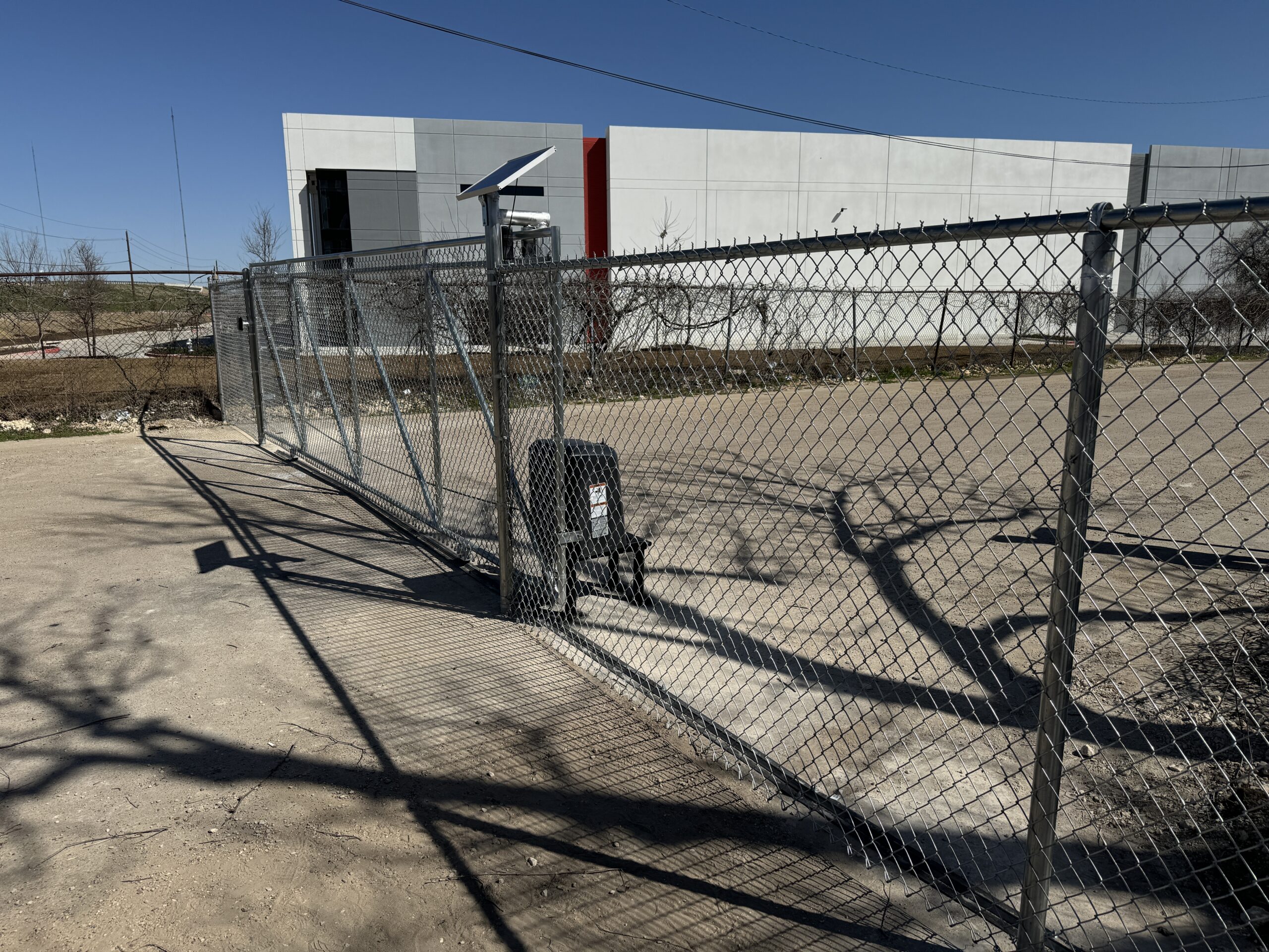 A chain-link fence runs across a dirt lot, next to an iron fence installation site, with a solar-powered security camera on a post and a black power box on the fence. A large white and red industrial building stands in the background under a clear blue sky.