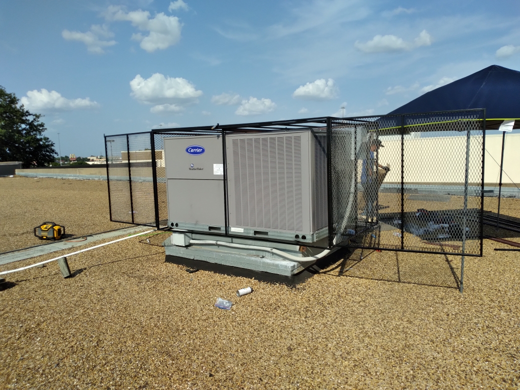 A large outdoor air conditioning unit, enclosed by a black iron fence installation, is set on a gravel-covered flat rooftop under a partly cloudy sky. Tools and debris are scattered nearby.